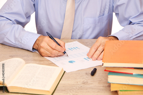 A businessman in a blue shirt and a beige tie in the office writes a report, on the table a globe, books and glasses.