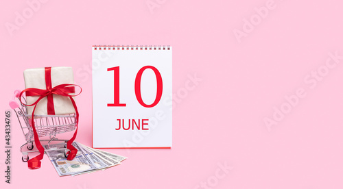 A gift box in a shopping trolley, dollars and a calendar with the date of 10 june on a pink background. photo