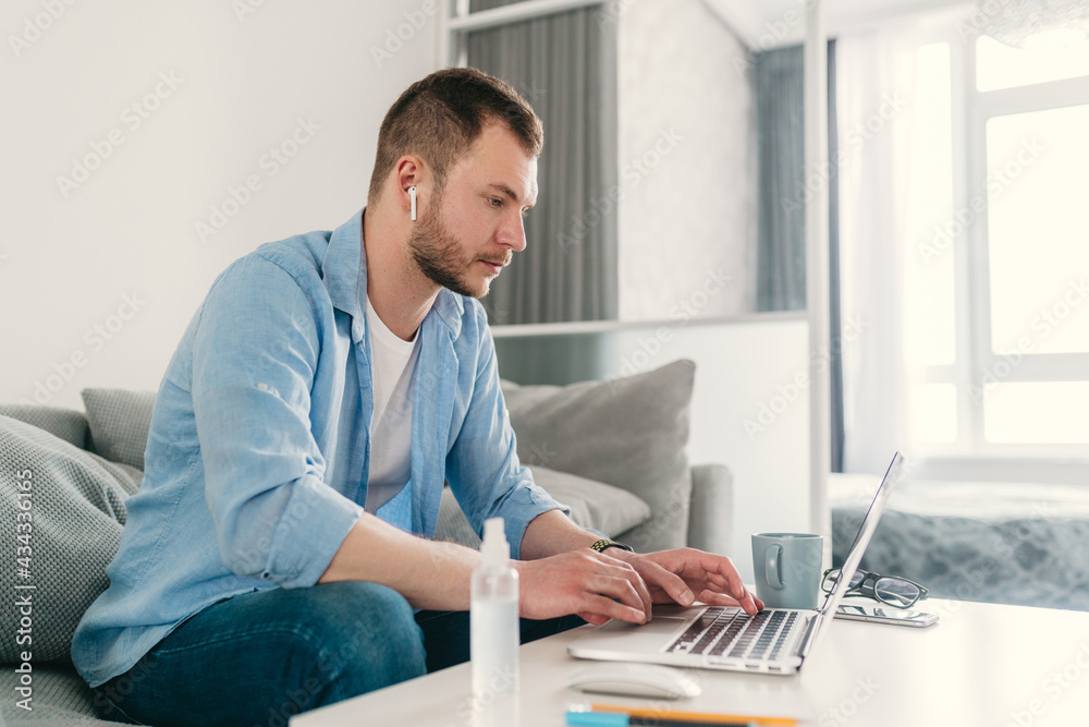 Fototapeta premium handsome smiling man in shirt sitting relaxed on sofa at home at table working online on laptop
