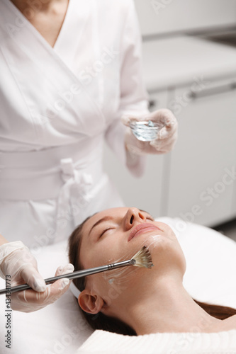 Fototapeta Naklejka Na Ścianę i Meble -  Beautician brushes ELOS gel with brush on beauty clinic patient, dermatologist using brush to insert cosmetic product on client face, preparaton for ELOS skin care treatment, remove facial redness