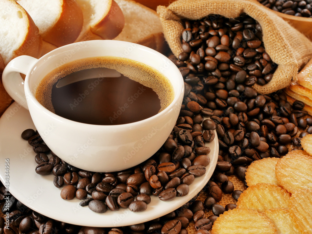 The white cup of hot dark coffee is between a sack of coffee beans and breads and circle crackers The white cup of hot dark coffee is between a sack of coffee beans and breads and circle crackers