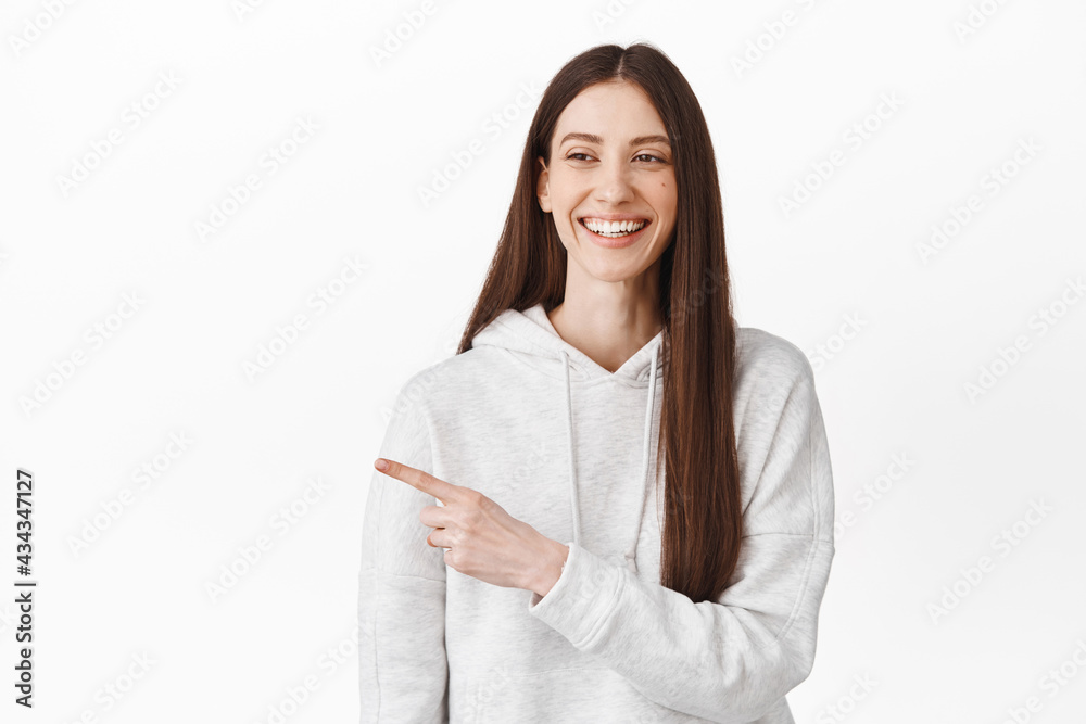 Cheerful young woman laughing, smiling and pointing aside, looking at logo with happy face expression, standing in casual clothes against white background Cheerful young woman laughing, smiling and pointing aside, looking at logo with happy face expression, standing in casual clothes against white background