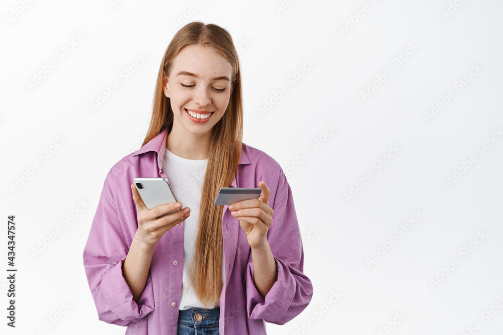 Smiling blond girl paying online, shopping in smartphone app and looking at her credit card, standing against white background Smiling blond girl paying online, shopping in smartphone app and looking at her credit card, standing against white background