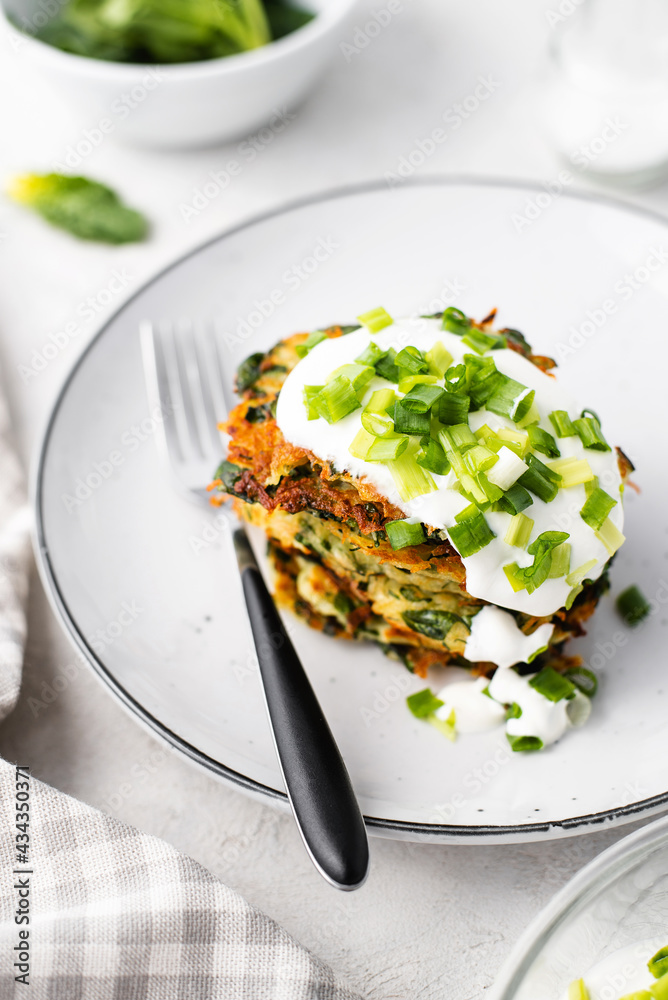 Potato pancakes with spinach, garlic and green onions served with sour cream dip on a plate on a light background Potato pancakes with spinach, garlic and green onions served with sour cream dip on a plate on a light background