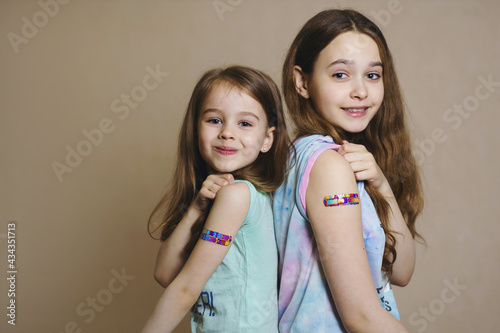 Two brave girls vaccinated against the coronavirus smile and roll up their sleeves to point to the vaccination site. Adhesive plaster on hands after vaccination against COVID-19 infection