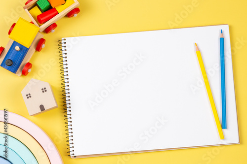 Blank sketchbook notebook with colored pencils and educational wooden kid toys and on yellow background. Top view. Early education, kindergarten, preschool, learn and play concept