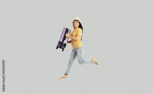 Holiday vacation and happy female tourist. Joyful excited dreaming young caucasian woman wearing casual clothes and summer hat jumping holding suitcase luggage studio shot on grey copy space