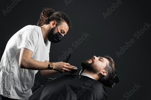 Beard shaving in barbershop. Barber with dreadlocks in black medical mask trim bearded man at quarantine coronavirus covid-19.