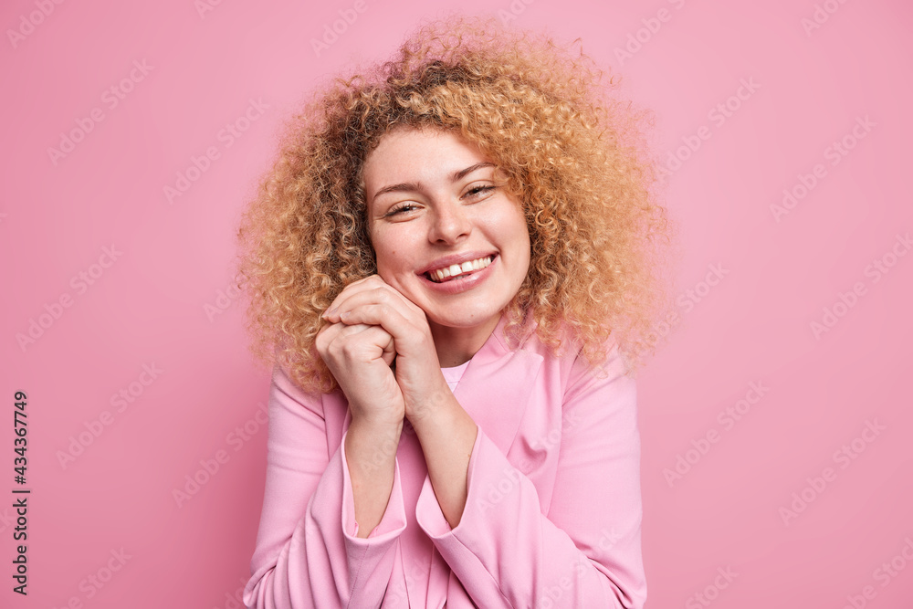 Portrait of happy sincere woman with curly hair keeps hands near face glad to hear heartwarming words or compliment smiles broadly dressed in pink jacket expresses positive emotions and feelings Portrait of happy sincere woman with curly hair keeps hands near face glad to hear heartwarming words or compliment smiles broadly dressed in pink jacket expresses positive emotions and feelings