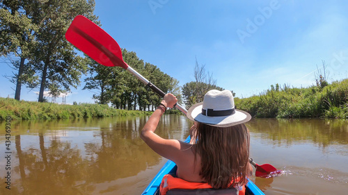 A woman in a white hat kayaking on Nida river in Poland. She is holding a red paddle in her hands. The banks of the river are overgrown with lush green grass. Clear, blue sky. Outdoor activity photo