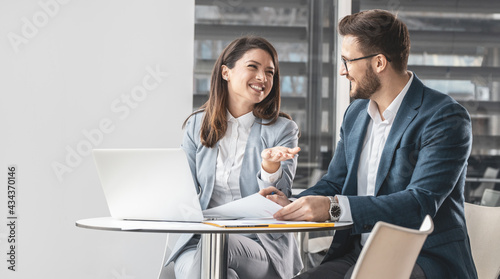 Two office workers discuss a new company project. They're sitting at the desk in the company cafe and preparing for a meeting.	 photo
