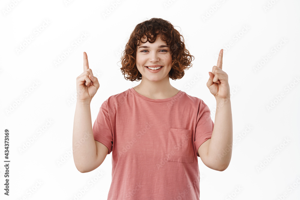 Image of smiling attractive woman pointing fingers up, showing top advertisement, grinning with happy and pleased face, introduce promo offer, standing against white background Image of smiling attractive woman pointing fingers up, showing top advertisement, grinning with happy and pleased face, introduce promo offer, standing against white background