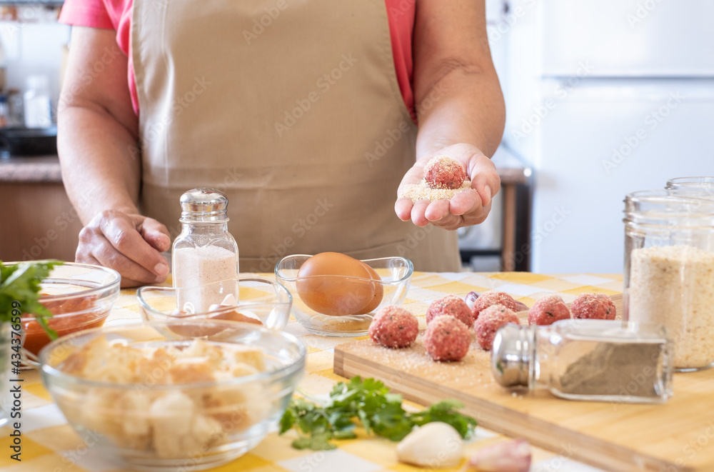 Woman's hand holding a fresh meatball ready to be cooked, wooden cutting board and raw ingredients