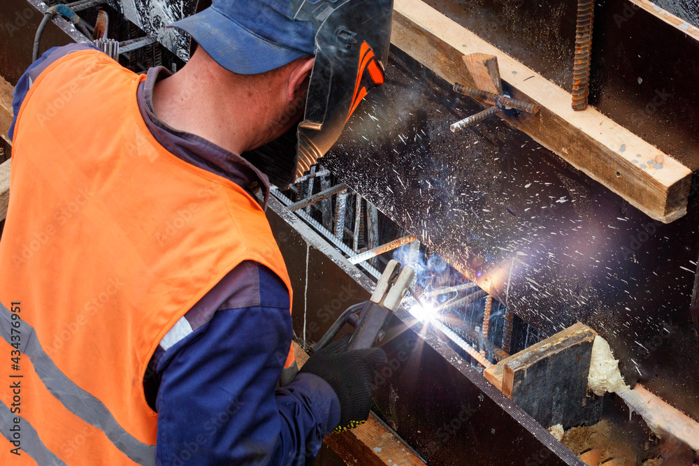 A welder in a protective helmet and gloves works with metal, welds metal rods during the construction of a wooden box for pouring a concrete structure.