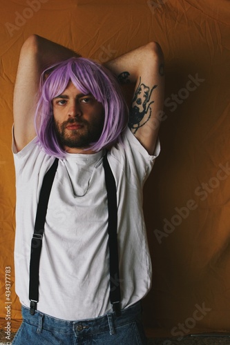 powerful portrait of queer young man with serious tough face expression with pink wig and tattoo on arm in orange background
