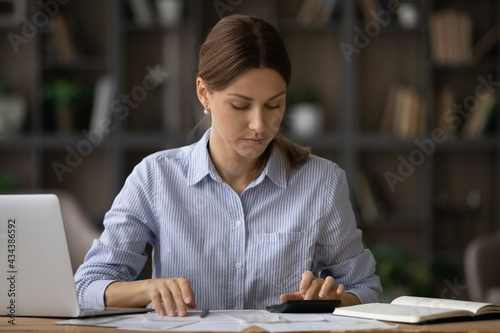 Serious millennial female social worker counting grant subsidy rates under government program at office workplace. Attentive young woman bookkeeper check accounts bills calculate company income taxes