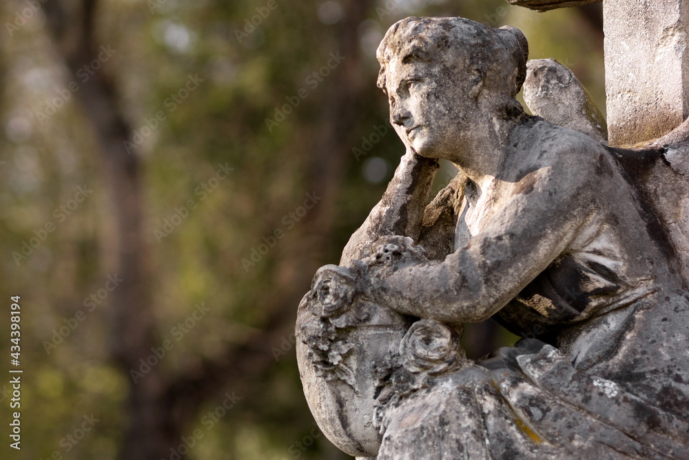 Beautiful sculpture of a seated angel with an illuminated face. Blurred trees in the background. Beautiful sculpture of a seated angel with an illuminated face. Blurred trees in the background.