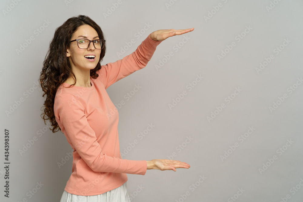 Excited woman hold open palm arms on empty space for promotion, sales and product presentation isolated over gray background. Emotional female in 30s with happy smile wear glasses present, studio shot Excited woman hold open palm arms on empty space for promotion, sales and product presentation isolated over gray background. Emotional female in 30s with happy smile wear glasses present, studio shot
