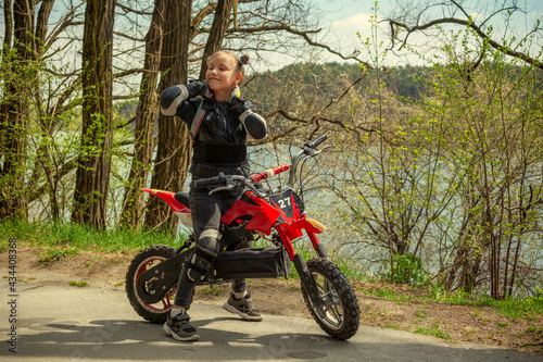 A child girl in motorcycle protection, knee pads and elbow pads on a cross motorcycle stands enjoying a walk on an asphalt path in the park near the river and trees in the spring