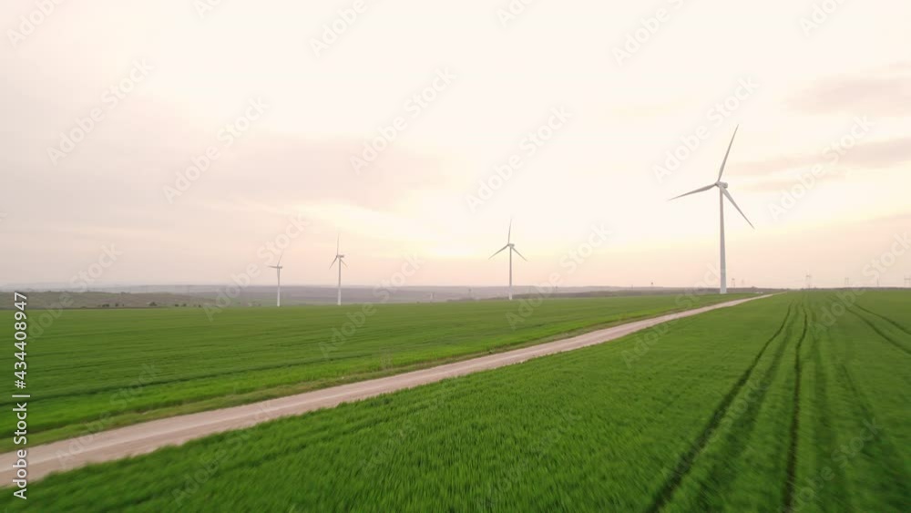 Car drives along rural road kicking up dust among green wheat field aerial view. Large wind turbines against blue sky at sunset in spring. Wind park agricultural farm. Alternative energy eco nature Car drives along rural road kicking up dust among green wheat field aerial view. Large wind turbines against blue sky at sunset in spring. Wind park agricultural farm. Alternative energy eco nature