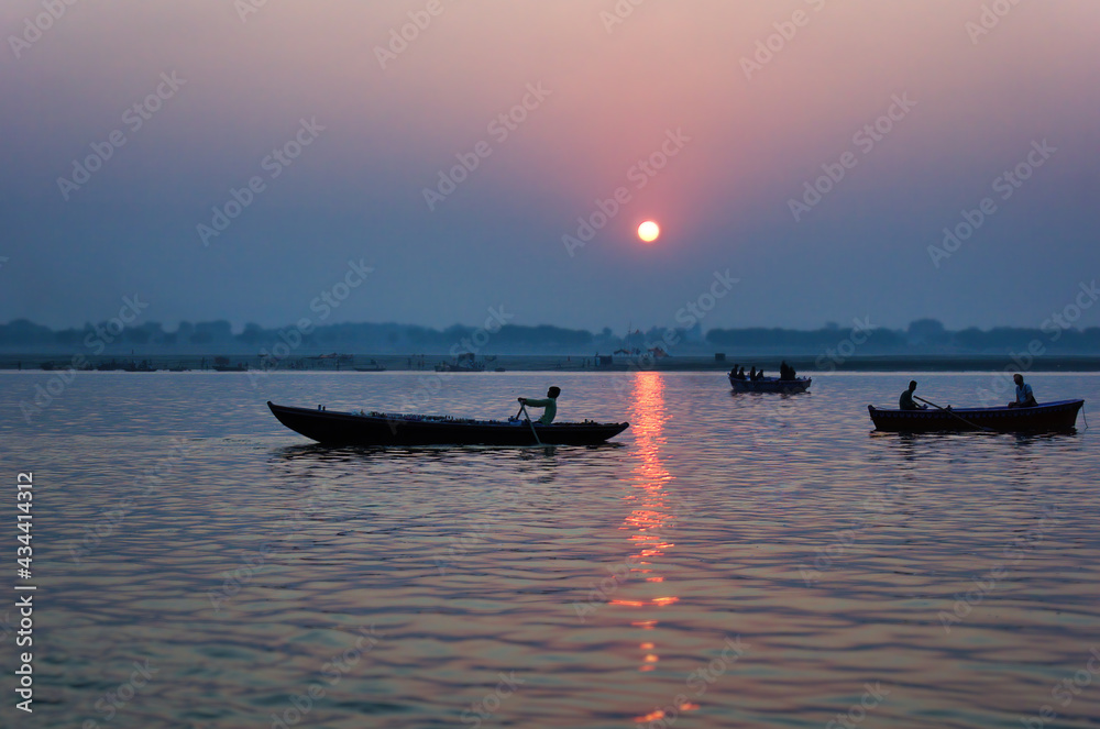Varanasi, India: Wide angle Silhouette shot of People and tourists on wooden boat sightseeing in ganges river close to famous Munshi ghat during sunrise or sunset.