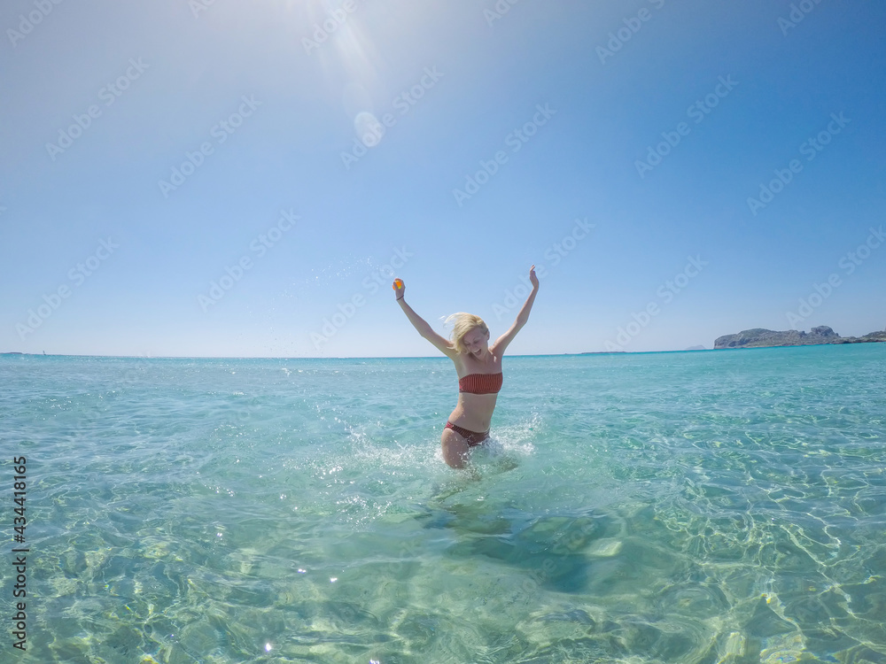 Cheerful woman in a bikini jumping and splashing water. She is in shallow turquoise water by the beach on Crete island. Cheerful woman in a bikini jumping and splashing water. She is in shallow turquoise water by the beach on Crete island.