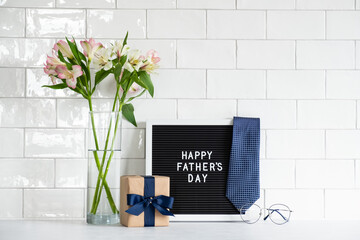 Happy Fathers day concept. Festive composition with gift box wrapped craft paper with blue ribbon, letterboard with text Happy Father's Day, necktie, boquet of flowers, glasses.