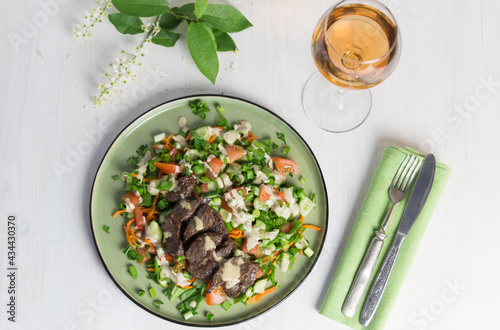 salad with fried beef  vegetables  sauce on green plate on white background  rose wine glass  bird cherry blooming branch  fork and knife on textile