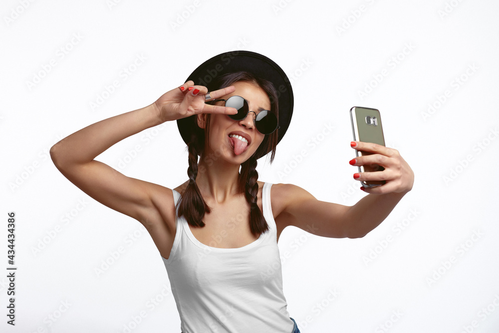 Cute girl taking selfie and show tongue and peace gesture over white wall