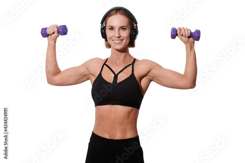 Strong muscular sportswoman in headphones and black outfit doing exercise with dumbbells over white isolated background