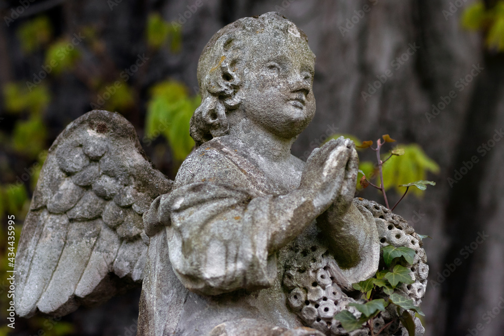 Sculpture of an angel in the cemetery. 
Blurry background with trees and green leaves.