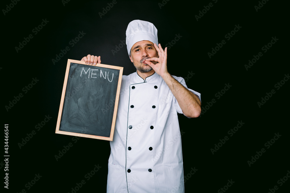 Chef cook wearing kitchen jacket and hat, holds a piazarra with the menu and makes good food gesture, on black background Chef cook wearing kitchen jacket and hat, holds a piazarra with the menu and makes good food gesture, on black background
