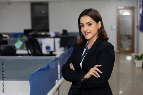 Portrait of young successful Indian business woman in black suit standing against office background with her arms folded.