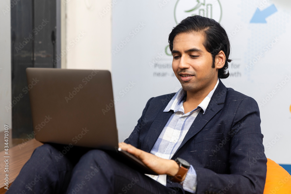 Portrait of young successful Indian business man in dark suit sitting and working on laptop, Corporate environment, expressive face. Portrait of young successful Indian business man in dark suit sitting and working on laptop, Corporate environment, expressive face.