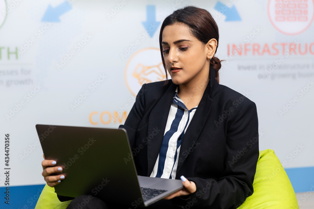 Portrait of young successful Indian business woman in black suit sitting on a bean bag and working on laptop, Corporate environment, expressive face. Portrait of young successful Indian business woman in black suit sitting on a bean bag and working on laptop, Corporate environment, expressive face.