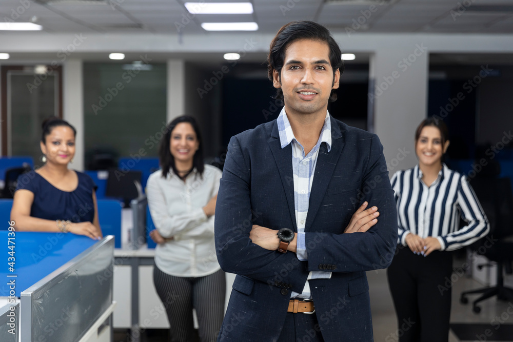Confident Indian businessman standing infront of his office colleagues, selective focus, corporate environment, team members, business office. Confident Indian businessman standing infront of his office colleagues, selective focus, corporate environment, team members, business office.