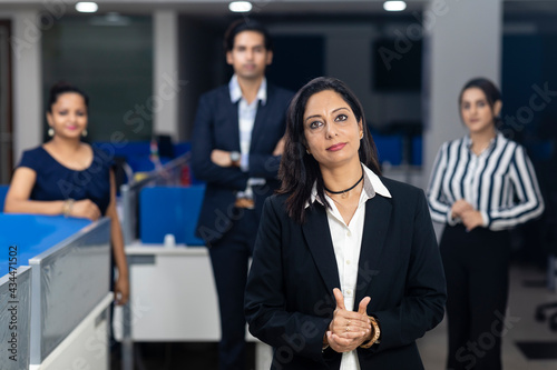 Confident Indian businesswoman standing infront of her office colleagues, selective focus, corporate environment, team members, business office.