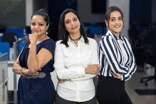 Three Indian businesswomen with arms folded standing together against office background, corporate environment.