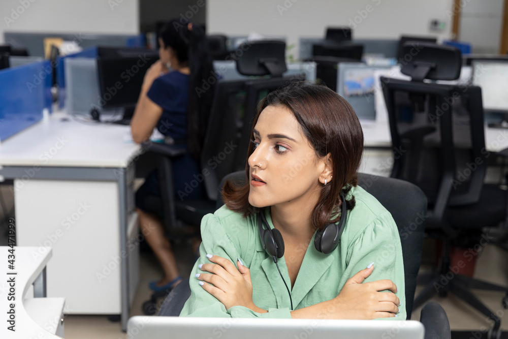 Portrait of pretty Indian businesswoman wearing headphones, sitting at her workstation against office background, corporate environment. Portrait of pretty Indian businesswoman wearing headphones, sitting at her workstation against office background, corporate environment.