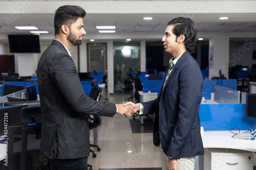 Portrait of two young Indian businessmen shaking hands, reaching agreement with client, closing a deal, corporate environment, office background.