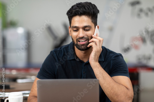 Portrait of a young handsome Indian man speaking on his phone and working on his laptop, sitting in an office cafeteria, coffee shop, casual work.