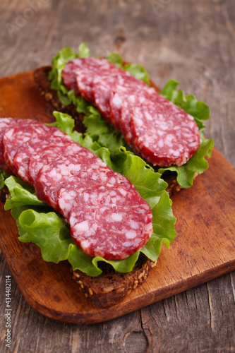 Delicious sandwich on black grain bread with lettuce and smoked sausage on old wooden background.