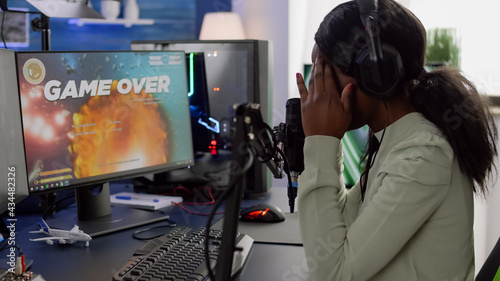 Furious black woman gamer with headphones using stream chat losing game playing on RGB powerful computer shooter game. Virtual online streaming cyber checking sound performing in gaming tournament