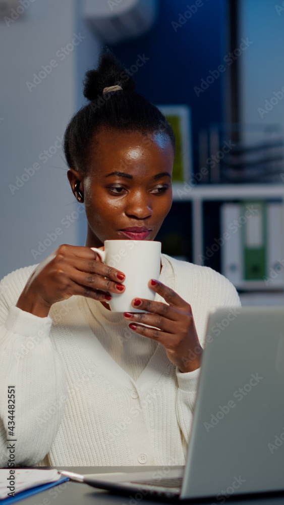 Black business woman using wireless headphone drinking coffee duing video conference working overtime from start-up office in front of laptop. Freelancer using talking on virtual meeting at midnight Black business woman using wireless headphone drinking coffee duing video conference working overtime from start-up office in front of laptop. Freelancer using talking on virtual meeting at midnight
