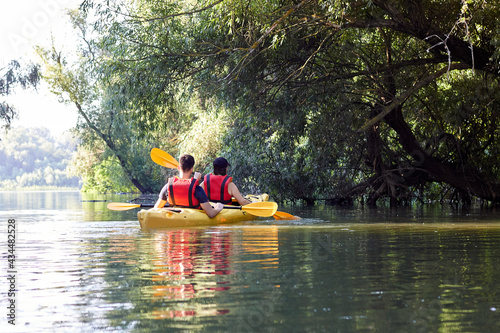 Mother and big son paddle yellow kayak near green trees on wildlife areas of Danube river. Summer kayaking. Concept for family, adventure, travel, action, lifestyle and kayaking