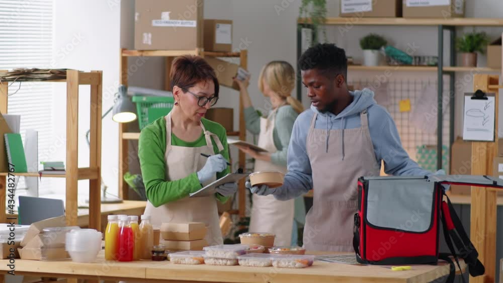 Caucasian female manager talking notes on clipboard and giving instructions to Afro-American male courier as he packing kraft paper lunch boxes with meal in food delivery bag