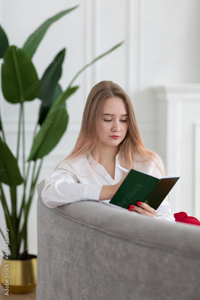 Blonde long haired young Caucasian business woman writing in planner notepad, diary, planning work and affairs schedule, sitting on sofa, vertical shot. Blonde long haired young Caucasian business woman writing in planner notepad, diary, planning work and affairs schedule, sitting on sofa, vertical shot.