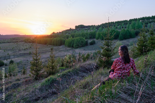 A brunette girl in a pink blouse sits on a green hill and looks at the sunset. Summer view, beautiful sky.
