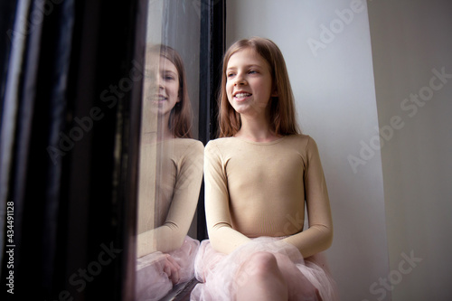 Beautiful teenage girl with long blonde hair in bodysuit and a fluffy tulle pink skirt sitting by the window. Girl dreaming