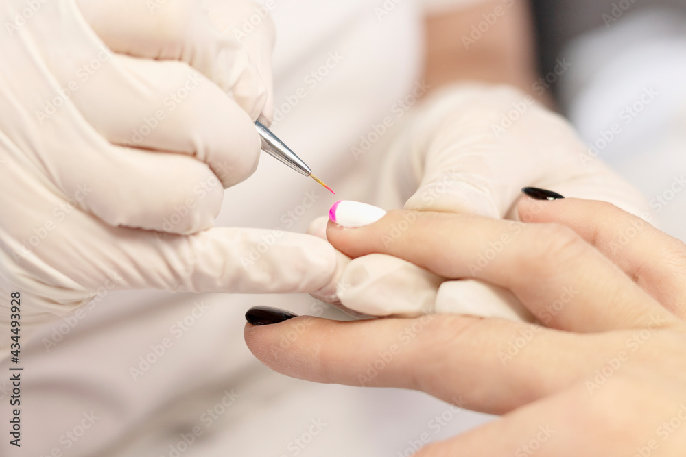 Manicure in a beauty salon. A manicure master draws a French jacket with a thin brush by hand on the nails of a young girl with gel polish. Space for the text. The concept of professional care for han Manicure in a beauty salon. A manicure master draws a French jacket with a thin brush by hand on the nails of a young girl with gel polish. Space for the text. The concept of professional care for han